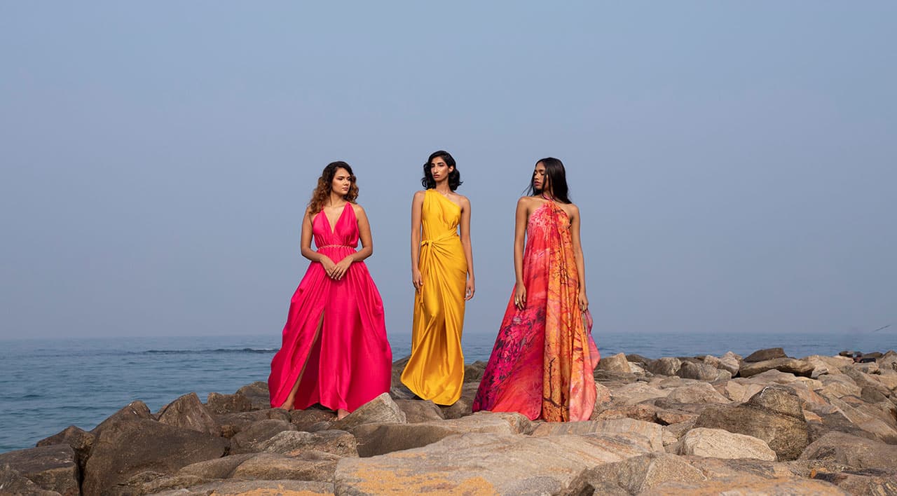 Three models in flowing gowns standing on coastal rocks by the ocean